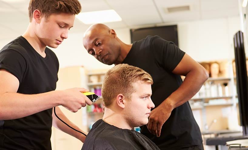 A teenage boy is buzz cutting a man’s hair while his teacher assists him. All three of them wear black shirts.