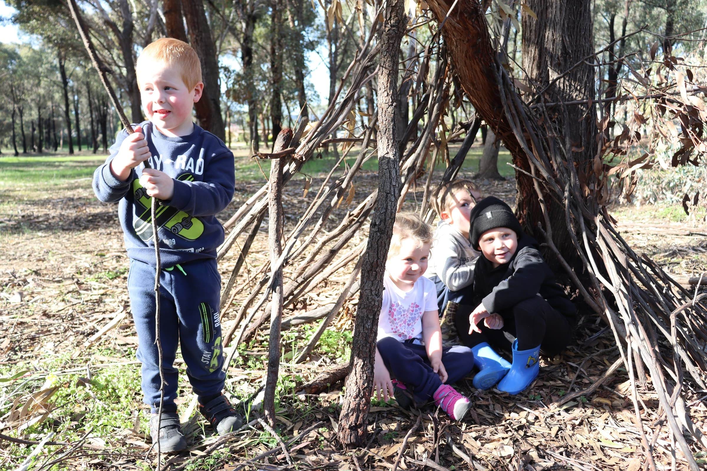 Three young children sitting under a tree in the bush with a young boy holding a stick.