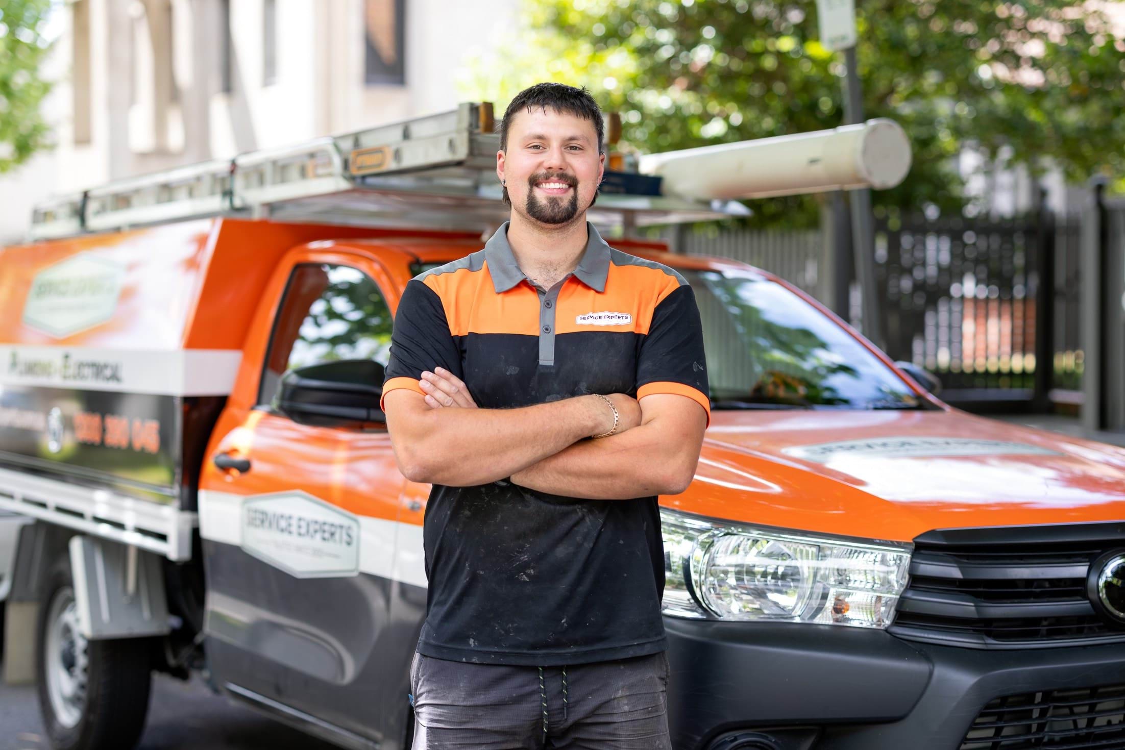 Tom is standing outside smiling with arms folded in front of a ute.
