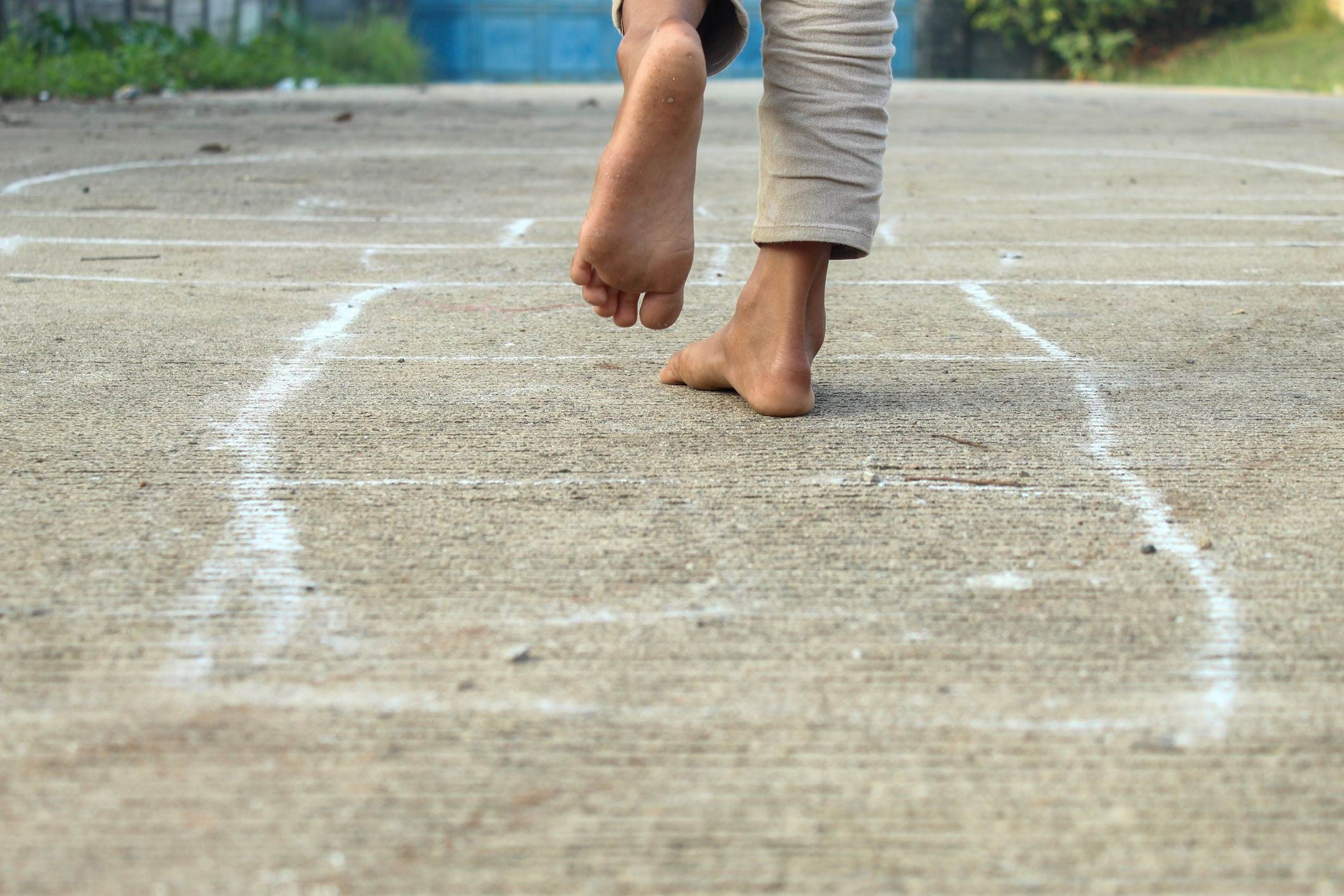 A child playing hopscotch outdoors. Their bare feet are seen jumping over a chalk line drawn on a cement road.